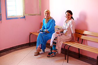 2 Moroccan women are sitting on bench and smiling at a rural health clinic